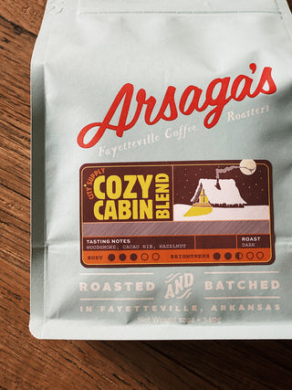 A light blue bag of coffee lays on a wooden surface. The bag showcases the brand name, along with the blend and the blend's notes.