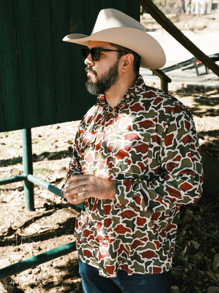 A man wearing an Amigo Provisions Razorback Camo Long Sleeve Button-Up Shirt, paired with a cowboy hat. The shirt features a unique camo pattern that incorporates the shape of the state of Arkansas within the camo segments. The shirt is buttoned up and fits the man’s frame, blending outdoor style with a touch of local pride.