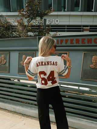 A woman outside of a football stadium showcases a cream t-shirt with grey lines around the sleeves, imitating a jersey, with the name being "Arkansas" and the number, "64" in red.