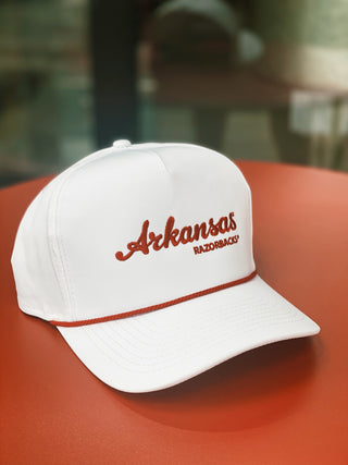 A white hat with a red rope with the words "Arkansas Razorbacks" in red threading, sits on a red table.