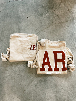 Two sweathirts lay folded on a concrete floor. One is the front of a cream sweatshirt with the red letters "AR" in the top left corner. The other is the back of a cream colored sweatshirt with the bolded red letters, "AR" centered with smaller white wording, "EST. 1871".