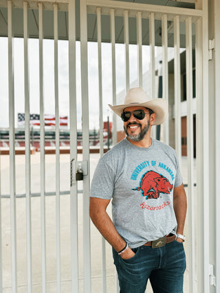 A man standing outside a football stadium wearing a grey t-shirt that features a vintage razorback logo, with the words "University of Arkansas" in blue, above the razorback; and "Razorbacks" in red, below the razorback.