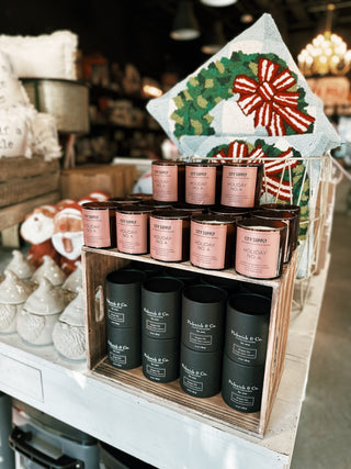 A Christmas table showcases many different candles. The top of the display a dark colored glass candle with a pink label that reads "City Supply Holiday No.4"