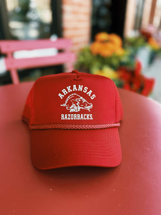 A red hat sits on a red table outside. The Hat features the words, "Arkansas" arched above a vintage inspired Razorback logo, and Razorbacks straight across below the logo. The hat as a rope running a top the slightly curved bill.