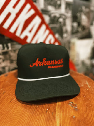 A black hat with a white rop and the words "Arkansas Razorbacks" in red threading, sits on a wooden surface.