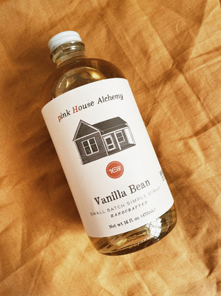 "A bottle of Pink House Alchemy syrup in the flavor 'Vanilla Bean' neatly arranged on a fabric cloth. The lightly amber-colored syrup is visible through the clear glass bottle,  sealed with a white cap and labeled with a minimalist white design featuring the brand's logo and flavor name."