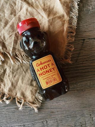 A bottle of Arkansas Hot Honey lying on a wooden surface. The clear bottle reveals the golden, slightly amber-colored honey inside, with a bold label featuring the product name and branding.