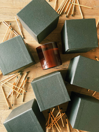 Square packages surround a dark amber glass candle, scattered around wooden matches.