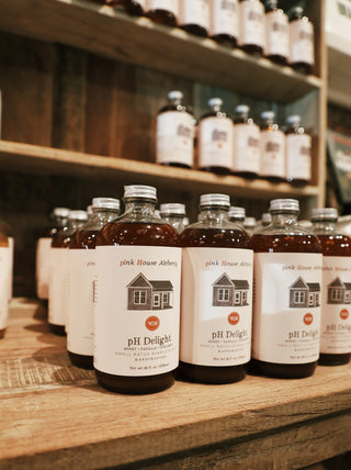 "Several bottles of Pink House Alchemy syrup in the flavor 'PH Delight' neatly arranged on a wooden surface. The amber-colored syrup is visible through the clear glass bottles, each sealed with a black cap and labeled with a minimalist white design featuring the brand's logo and flavor name."