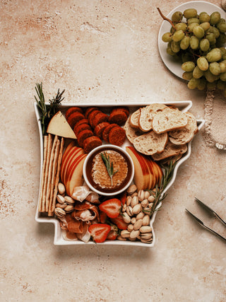 An Arkansas state-shaped white ceramic dish filled with a charcuterie mix, showcasing a potential use for the plate. The dish is arranged with a variety of meats, cheeses, crackers, and fruit, with the state outline clearly visible. The food is artfully arranged to highlight both the plate’s unique shape and its functionality as a serving tray.