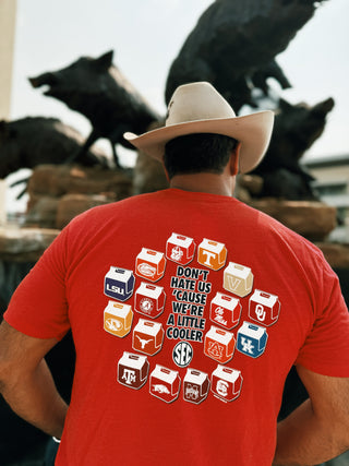 A man wearing a cowboy hat and a red t-shirt featuring logos of SEC schools on illustrated coolers. The shirt has the playful phrase 'Don't hate us cause we're a little cooler' in bold text, with the SEC logo prominently displayed. The design reflects a fun, spirited nod to college sports culture.