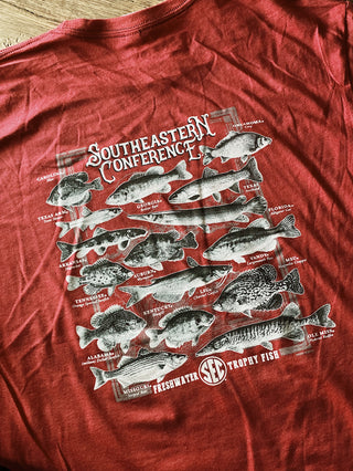 A red t-shirt laid on a wooden surface, proudly displays multiple different kinds of fish, assigned South Eastern Conference school teams. 