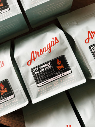 A light blue bag of Arsaga's coffee in the 'City Supply Campfire Blend' flavor sits on a wooden surface, accompanied by several other bags of the same flavor. The bags feature the distinctive branding and flavor name in bold letters.