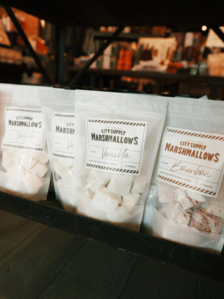 A package of City Supply Marshmallows in the flavor 'Vanilla' placed next to several other flavors of the marshmallows. The vanilla flavor is in a clear bag with a simple label showcasing the brand name and flavor. The other marshmallow bags, in various colors and designs, are visible beside it, highlighting the range of flavors available.